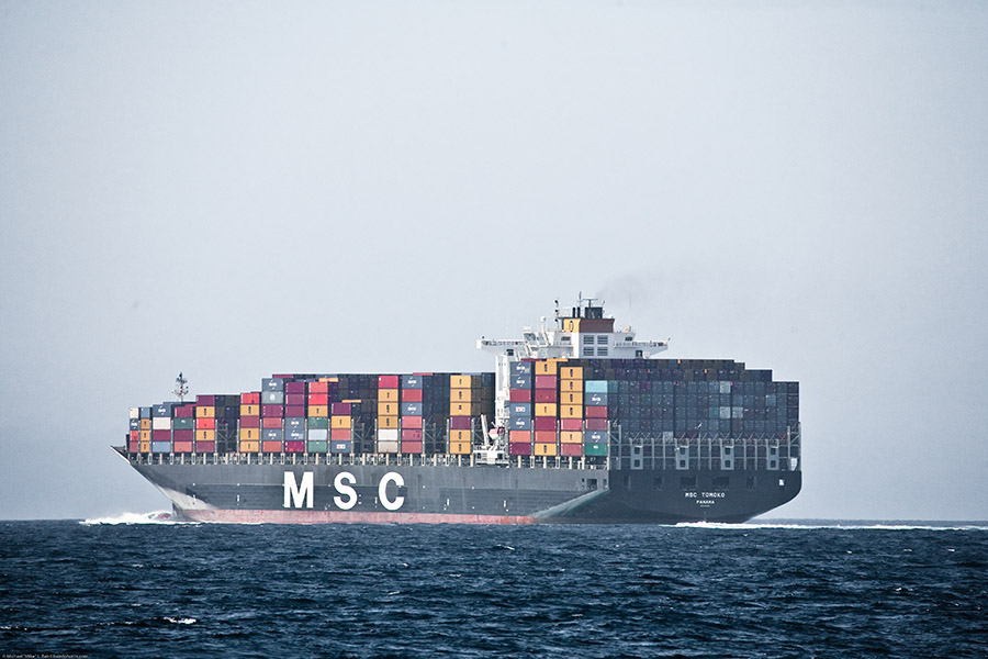 Cargo ship loaded with freight containers at sea with a muted, gray sky. (Photo Courtesy: Mike Baird via Flickr)
