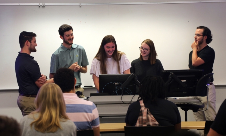 A group of students stand around a computer screen at the front of a classroom. 