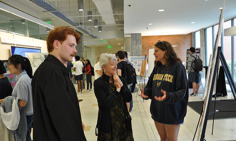 Woman gazing at art on easel being explained by a red-haired man on her left and a woman in a Georgia Tech sweat shirt on her right in the Mason lobby