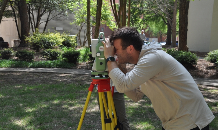 Male student in beige hoodie sighting into surveying equipment in a grassy area.