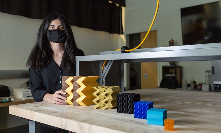 Larissa Novelino, a Ph.D. student at Georgia Tech, has long black hair and is wearing a black shirt and a black mask ovrer her nose and mouth. She is standing next to a table showing several metamaterial prototypes at different scales. They are different colors and range in size from smallest to largest.