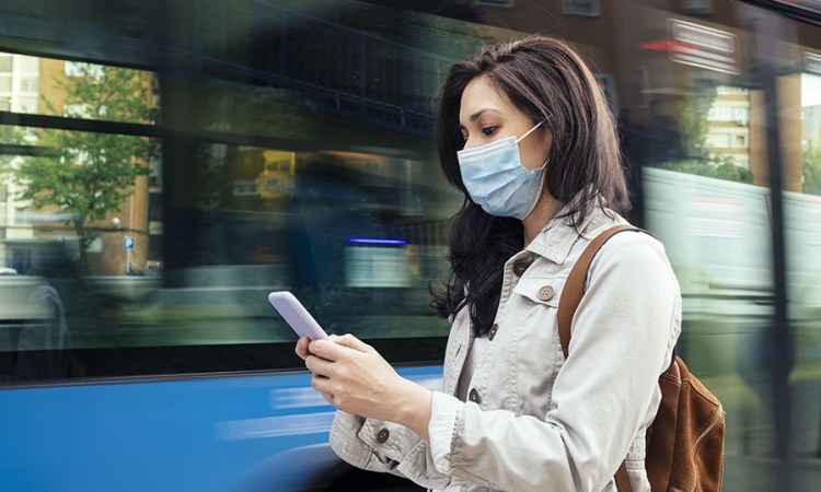 A woman wearing protective face mask looks at her smartphone as a bus goes by