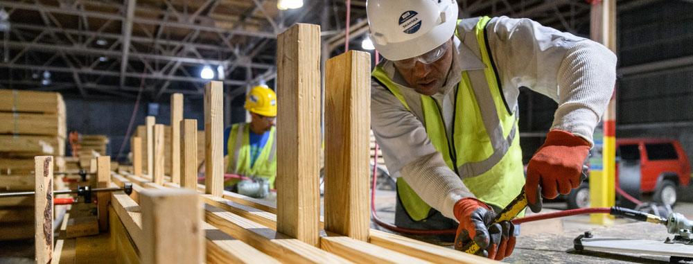 A man in a hard hat, gloves and vest cuts lumber