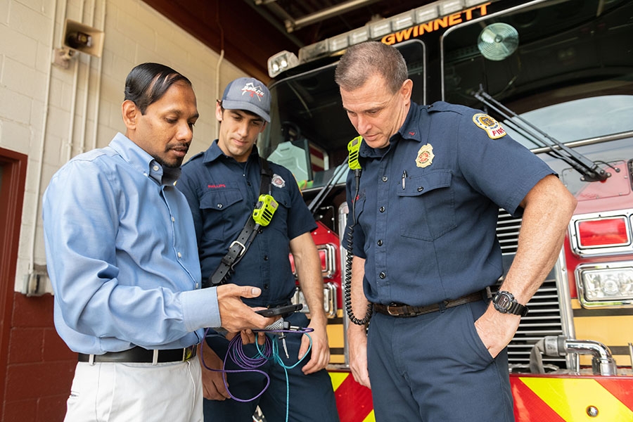 A man demonstrates a smartphone app for two firefighters standing in front of a fire engine