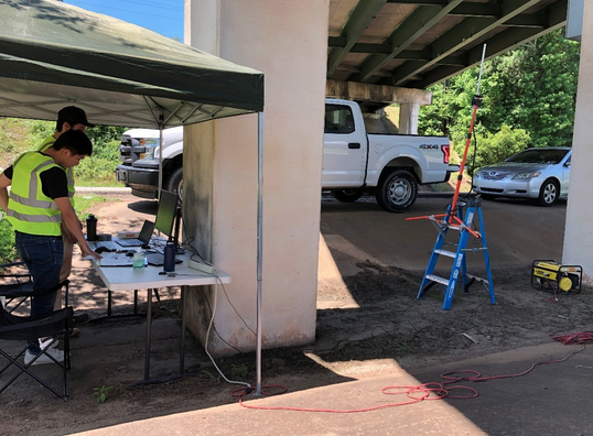 two people in neon protective vests look at a computer under a shade tent