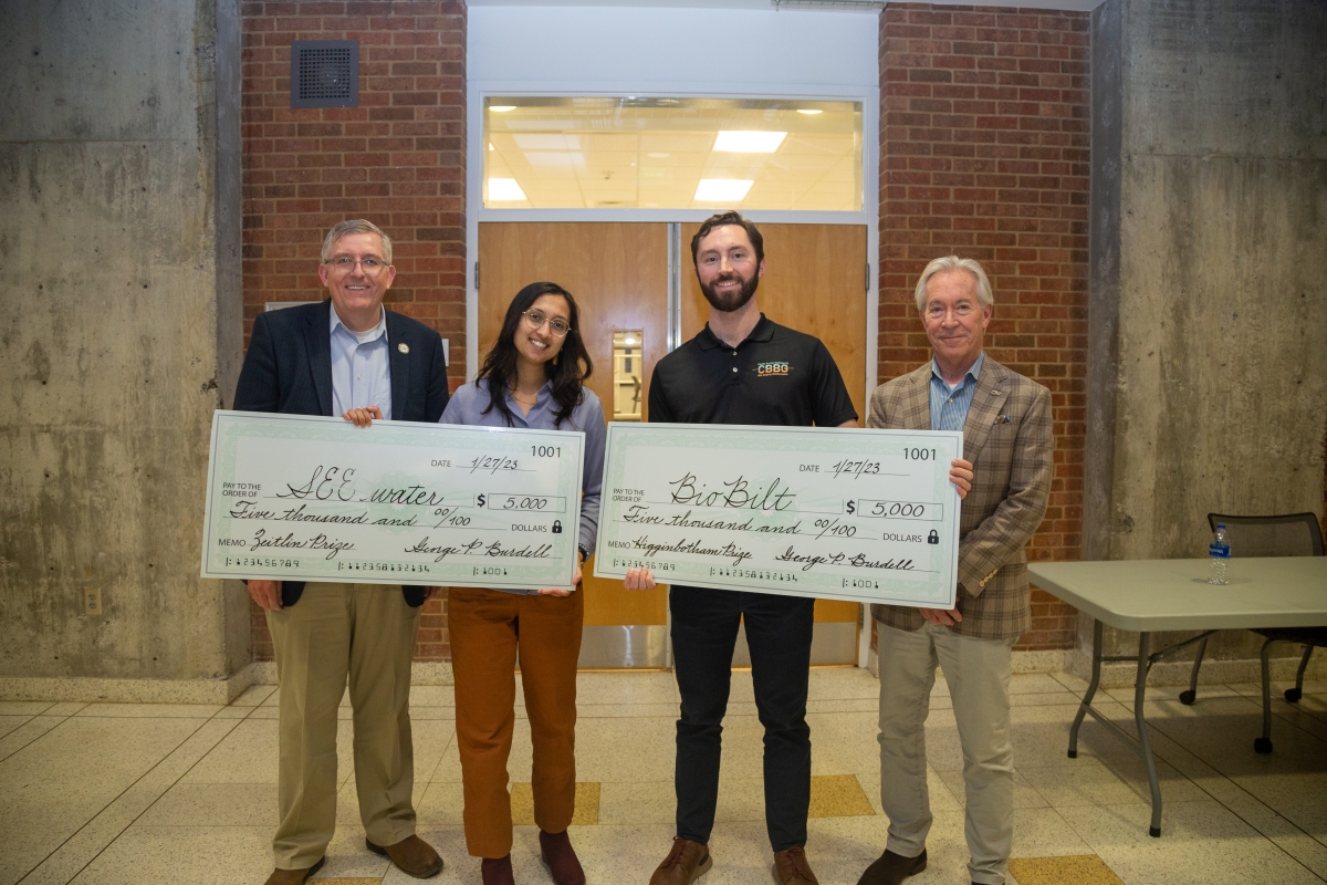 Four people pose holding two oversized checks 