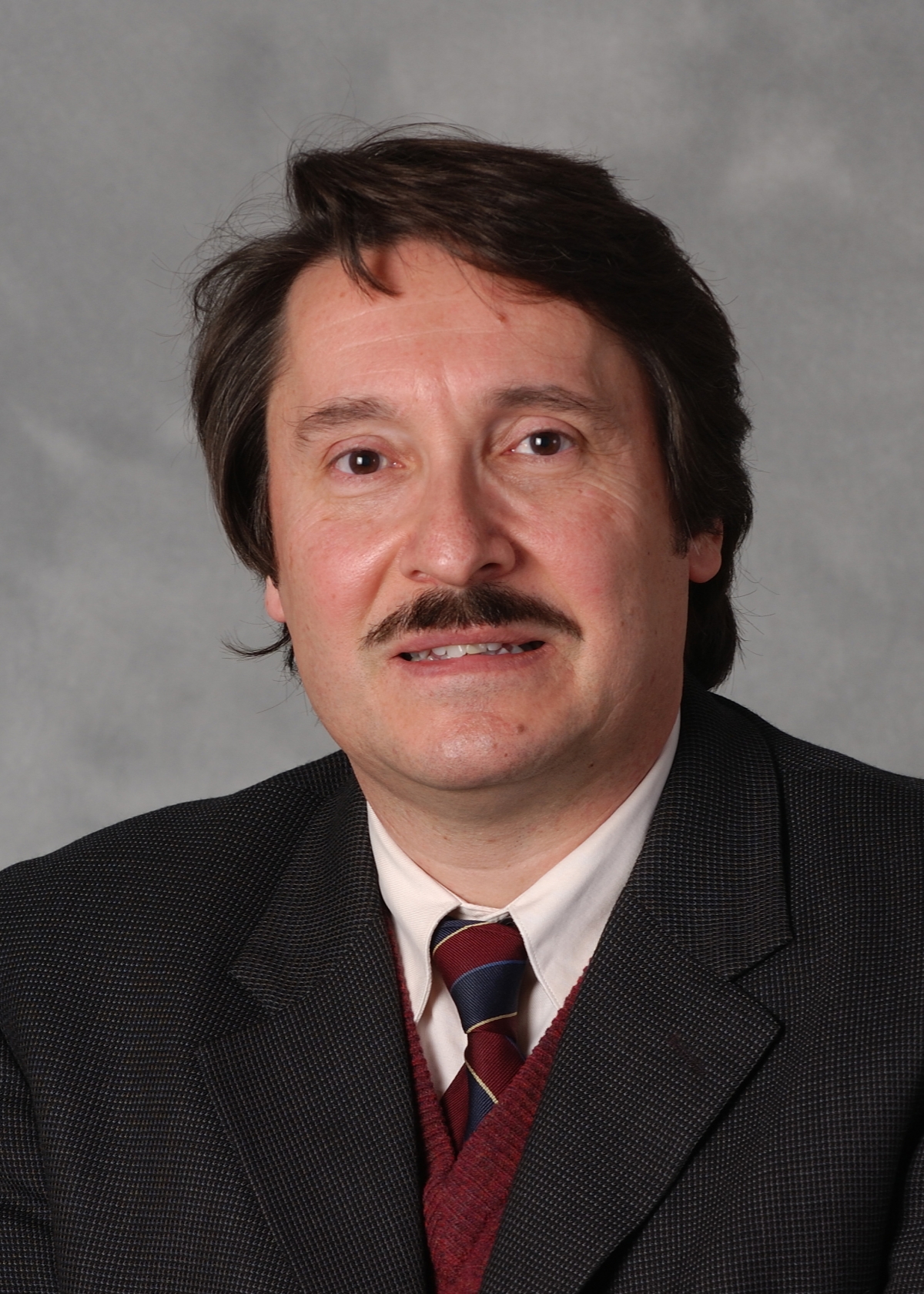 Headshot of a man in a dark grey suit, red vest, and patterned tie