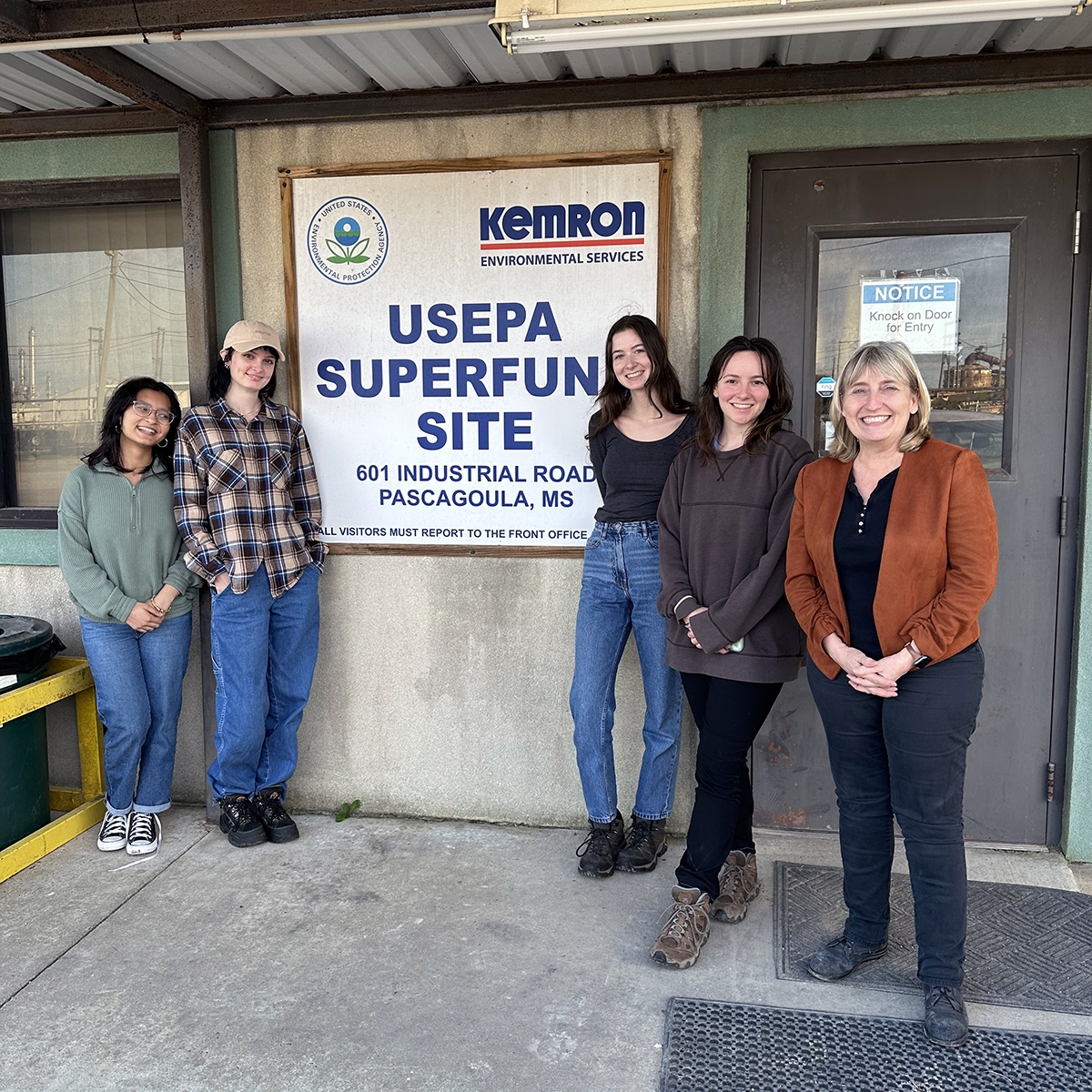 A group of four students stand with their professor outside a Superfund site office