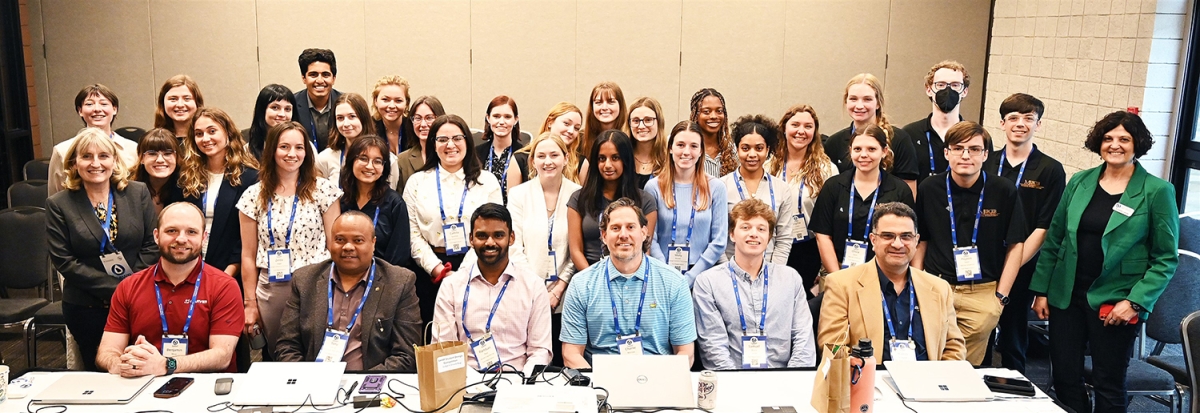 A group of student teams standing behind a seated panel of judges