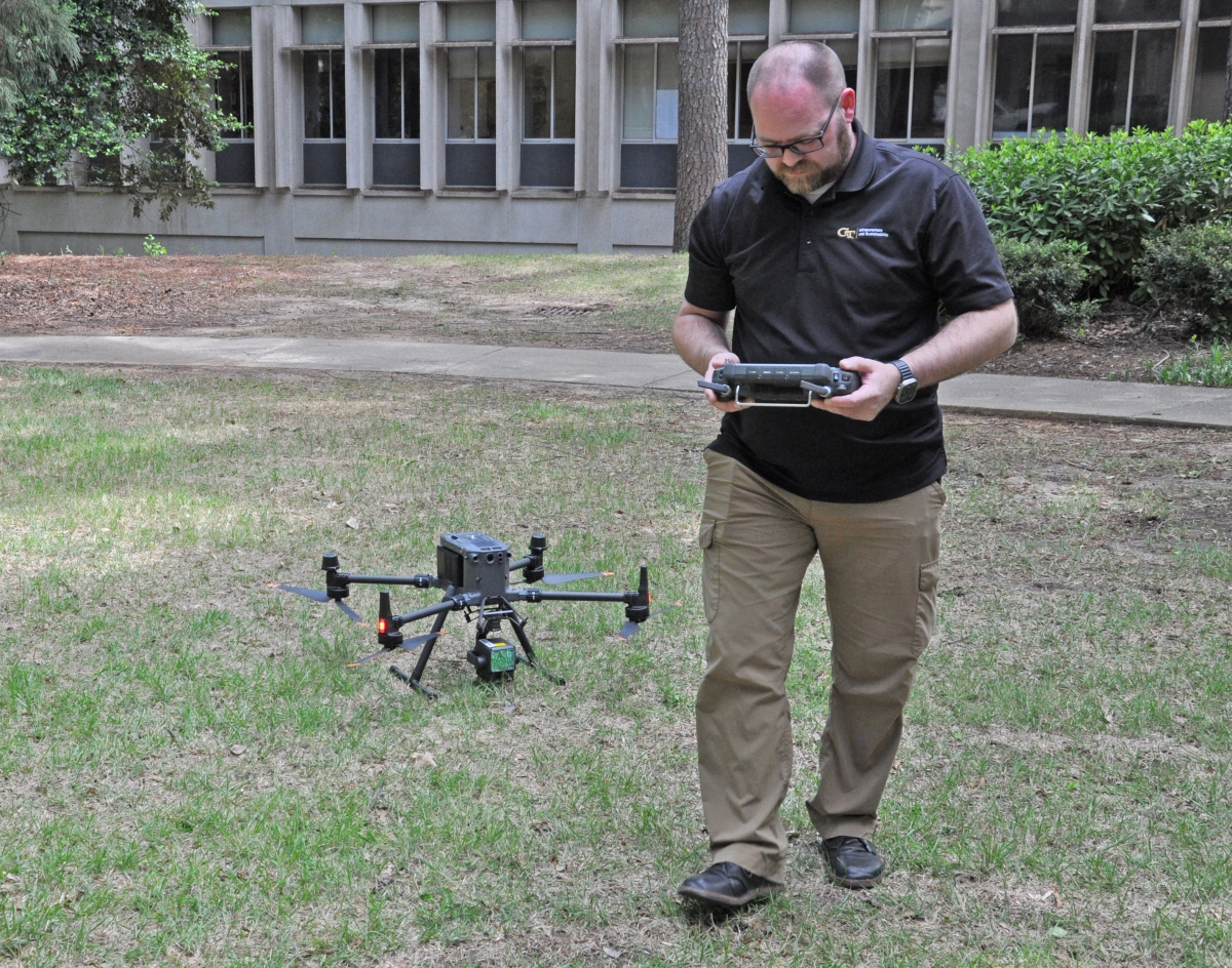 Man in a black Georgia Tech shirt and khaki pants holding the controller next to a drone on the ground next to him.