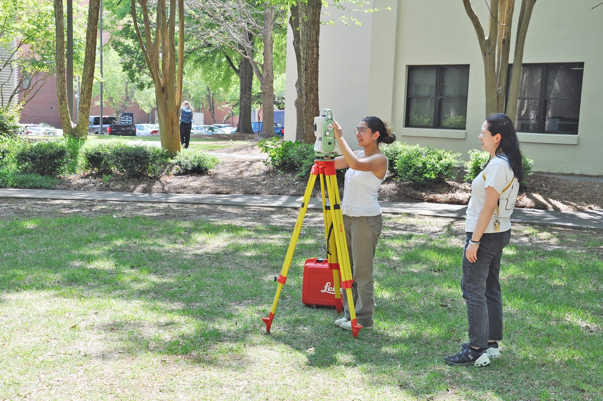 Female student in white top setting up surveying equipment while female graduate assistant looks on.