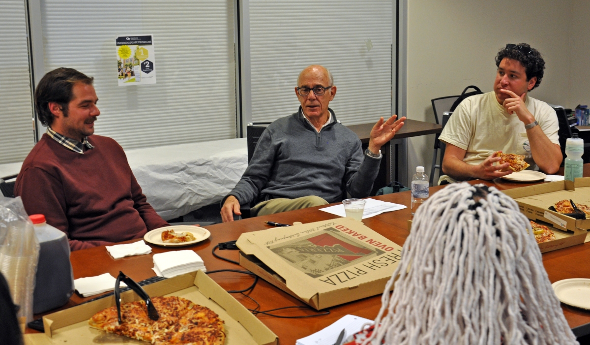 Dr. Raphael Bras gesturing seated at a conference table surrounded by students eating pizza