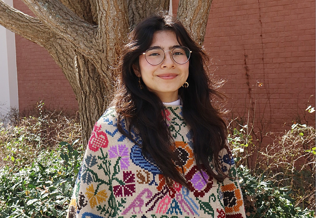 Ahtziri Rosales Ramirez posing in a knitted poncho in front of a tree on the Mason Building plaza