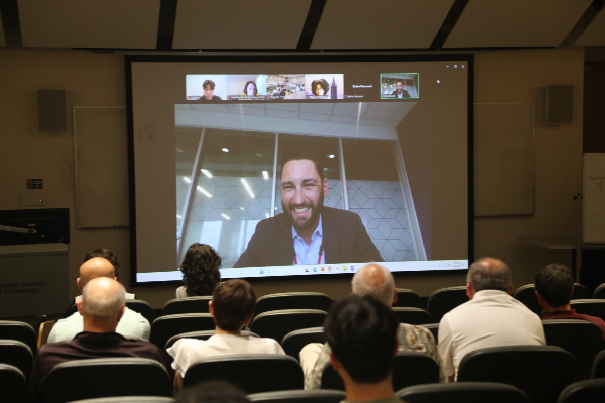 A man addresses a crowd on a screen at the front of a room 