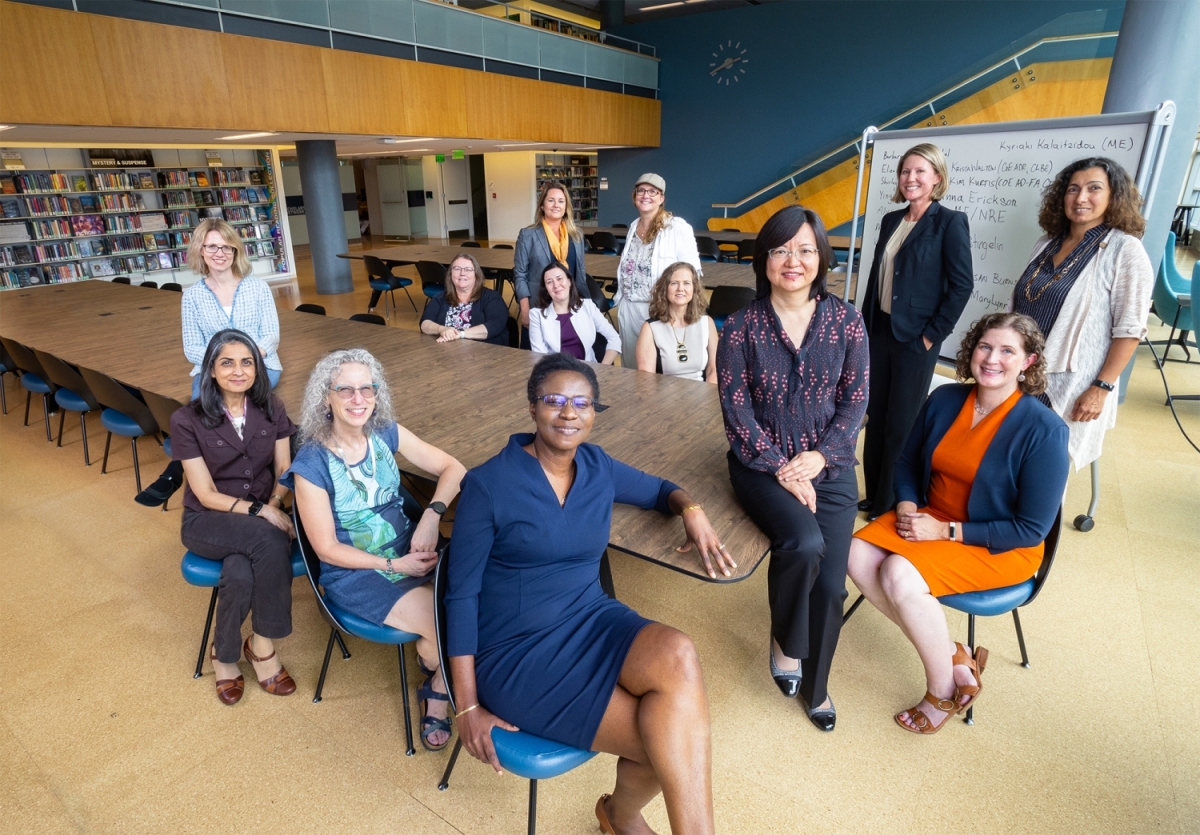 13 women in professional attire pose around a table in an academic space