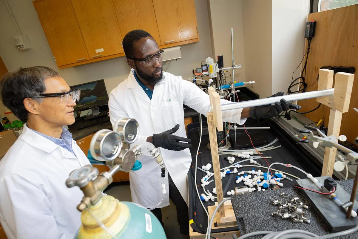 Two men in lab coats and glasses work with a reactor in a lab