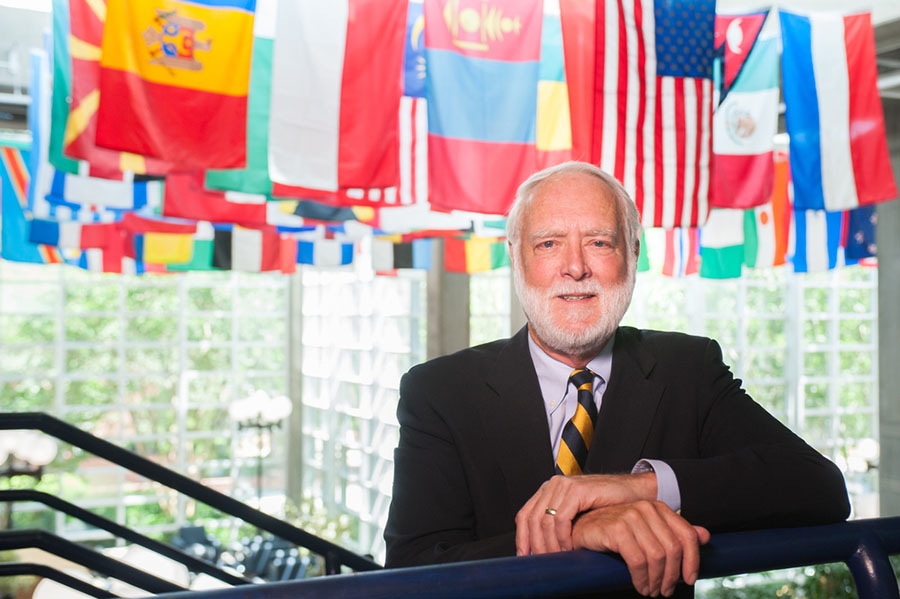 Georgia Tech President Emeritus G. Wayne Clough stands with his arms resting on a stair handrail with international flags hanging in the background.