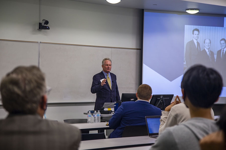 A man in a suit and tie gives a lecture with a screen to his right. The backs of people's heads in attendance can be seen in the foreground.