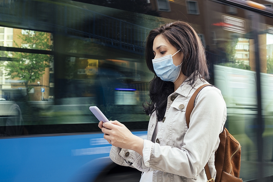 A woman wearing a blue surgical map looks at her smartphone as a bus drives by