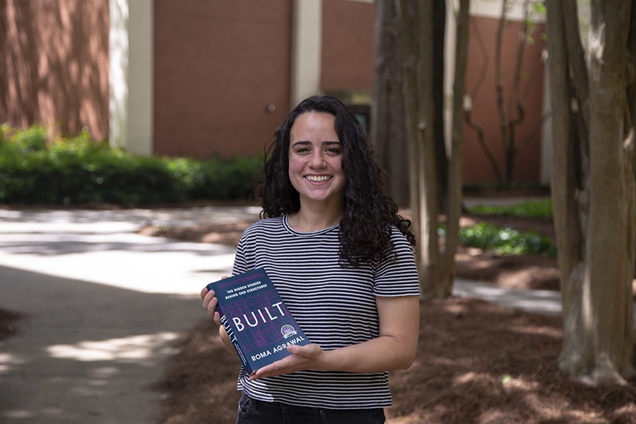 Maria Warren holds the book "BUILT" by Roma Agrawal standing in front of trees and a brick building.