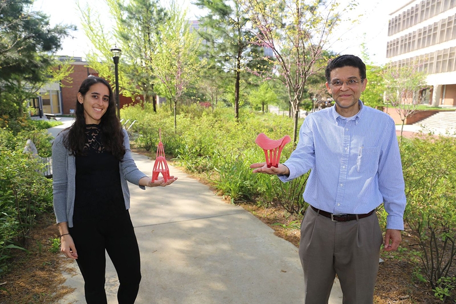 PhD Student Emily Sanders, left, and Professor Glaucio Paulino, right, hold red 3-D printed models as they stand on a campus sidewalk surrounded by greenery. 