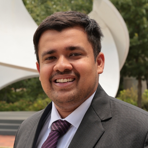 Portrait of Varun Desai in a white shirt with a burgundy striped tie and a grey blazer standing in front of the John Portman “Koan” statue